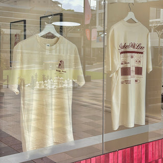 Two cream-colored graphic t-shirts displayed on hangers inside a store window. The left shirt features a small graphic with a tower and the text 'Ridge, Mass.