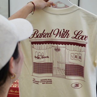 A cream-colored t-shirt with a dark red graphic print of a bakery storefront and the text 'Baked With Love' and 'Baby Buck'. A person wearing a white cap is holding the shirt on a hanger, with their hand and face partially visible.