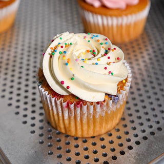 A close-up of a frosted cupcake with swirl of white icing and colorful sprinkles, sitting on a perforated metal tray with other cupcakes blurred in the background.