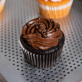 A chocolate cupcake with piped dark brown frosting and chocolate chips, sitting on a perforated gray metal tray with blurred cupcakes in the background.