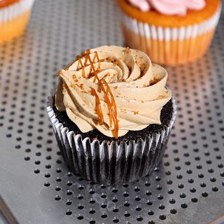 A single frosted cupcake in a black paper liner sits on a perforated metal tray. The cupcake has light tan whipped frosting with caramel drizzle and cookie crumbs, with other cupcakes blurred in the background.