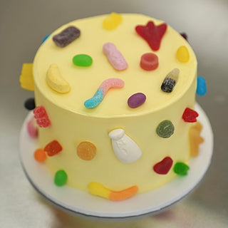 A round yellow frosted cake decorated with colorful gummy candies and sweets, sitting on a white plate against a neutral light gray background.