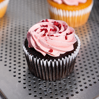 A chocolate cupcake in a white paper liner with light pink frosting and red berry pieces on top, sitting on a perforated metal tray with other cupcakes blurred in the background.