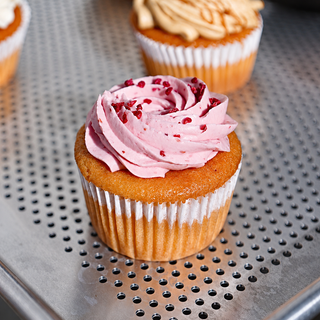 A close-up of a frosted cupcake with pink swirl frosting and red berry pieces, sitting on a perforated metal tray. Another cupcake is blurred in the background.