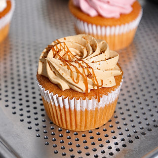 A close-up of a frosted cupcake with light tan swirl frosting, a white wrapper, and a drizzle of caramel sauce on a perforated metal tray, with another cupcake blurred in the background.
